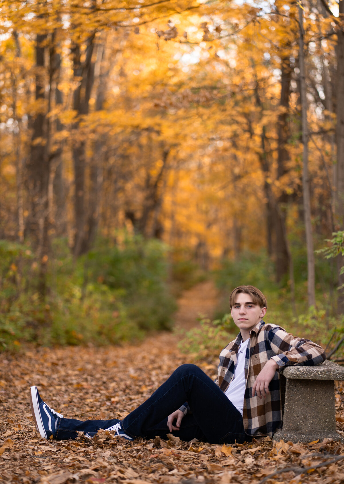 male teenager propped up against a stone bench on a wooded path with yellow leaves