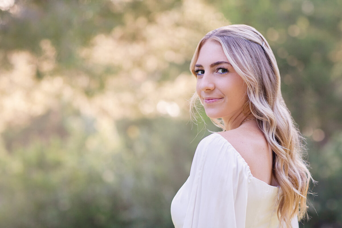 A woman with long blonde hair and a white off-shoulder dress stands outdoors, turning to glance back at the camera. The background is a soft blur of green foliage, suggesting a peaceful nature setting.