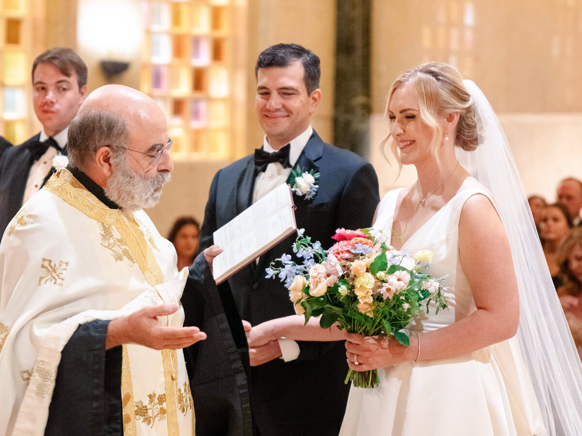 A bride and groom are smiling during their wedding ceremony. The bride holds a bouquet of colorful flowers, and a priest in a gold-embroidered robe is speaking to them, holding an open book. The setting appears to be a church with guests seated behind.