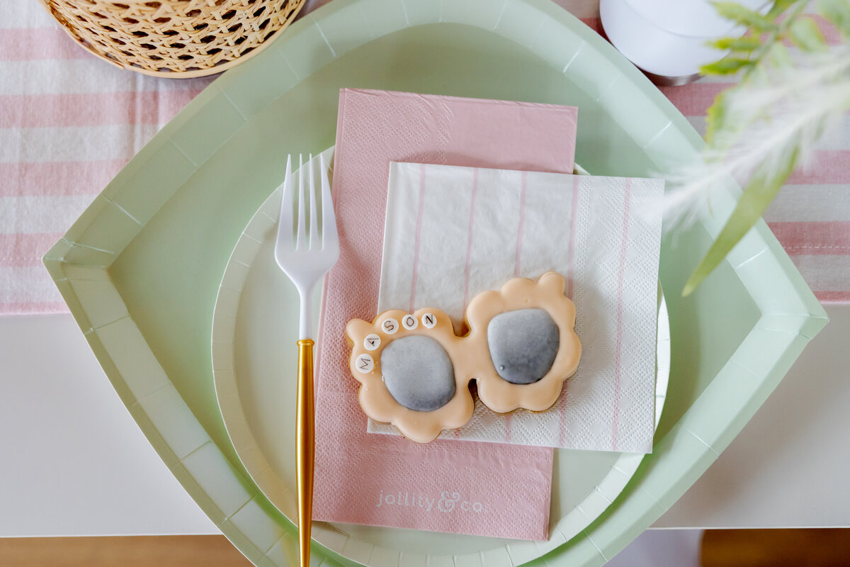 Kids table setting with cookie sunglasses, plastic gold utensils and pink and green plates and napkins.