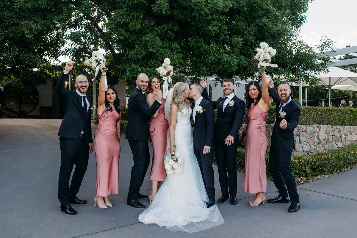 Wedding party dressed in pink and black cheering for the bride and groom who share a kiss in the centre. Outside Brindabella Hills Winery, Wallaroo NSW