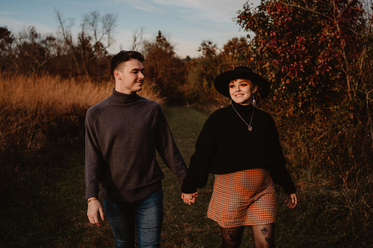 An expecting couple holding hands and walking on a grassy path 