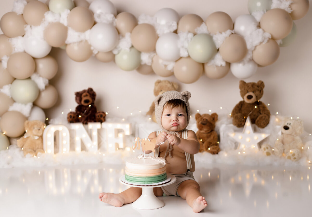 Teddy bear themed cake smash in West Palm Beach, FL photography studio. Baby's hands are full of cake, wearing a white teddy bear bonnet. Baby is staring at the camera. In the background there are white and beige balloons and stuffed teddy bears.
