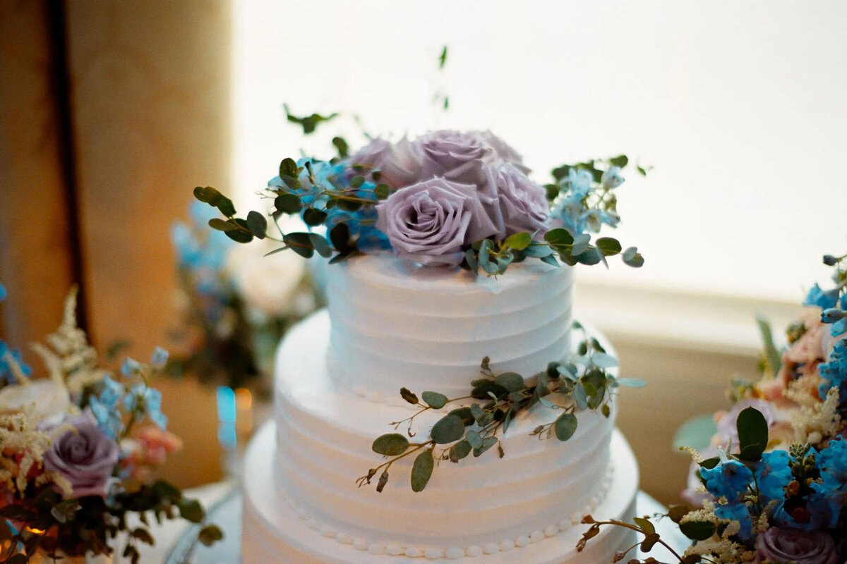 A white, two-tiered cake decorated with pale purple roses, blue flowers, and green leaves sits on a table against a softly lit background—perfect for an NJ wedding photographer to capture this elegant moment.