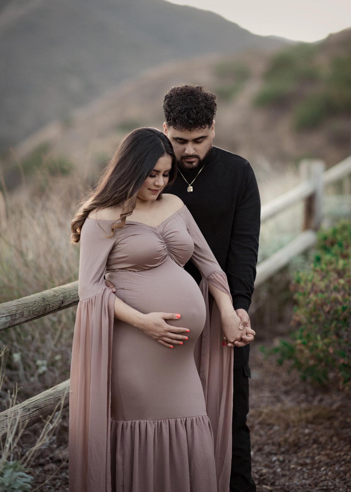 Expectant mom in long, beige maternity gown with side profile facing the camera, and Dad standing behind her facing the camera. Both are looking down a the baby bump, holding hands. Elsinore Mountains in the distance.