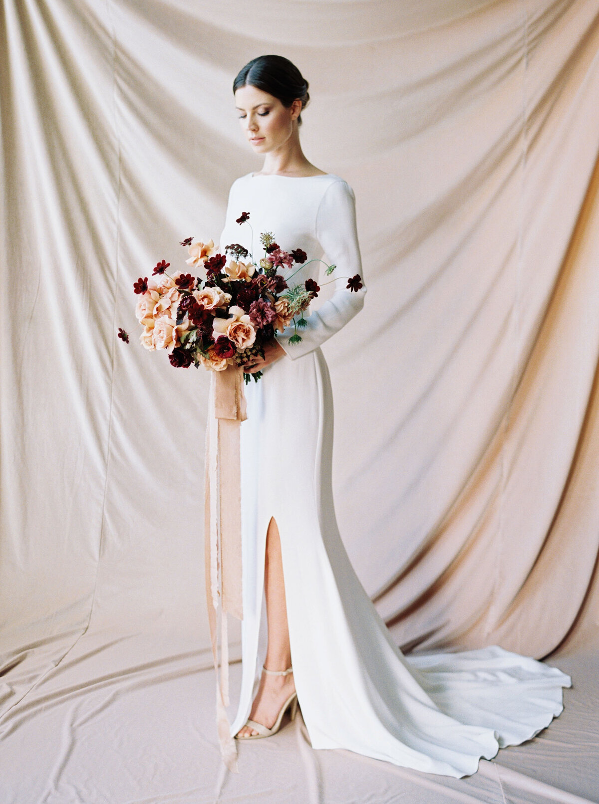 bride holding burgundy and peach floral boquet with peachy tone backdrop and white dress