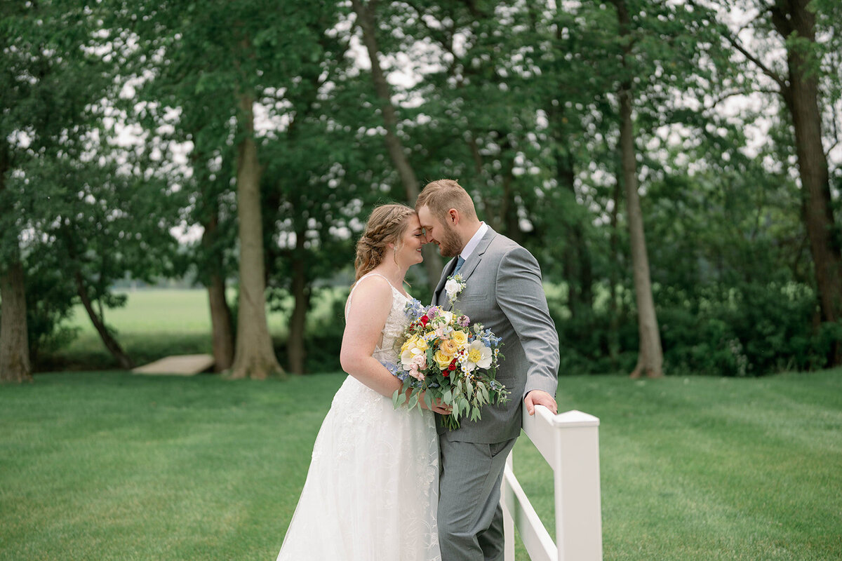 Bride and groom leaning against a white fence with their foreheads touching during wedding portraits at The Blue Heron Barn in Michigan.