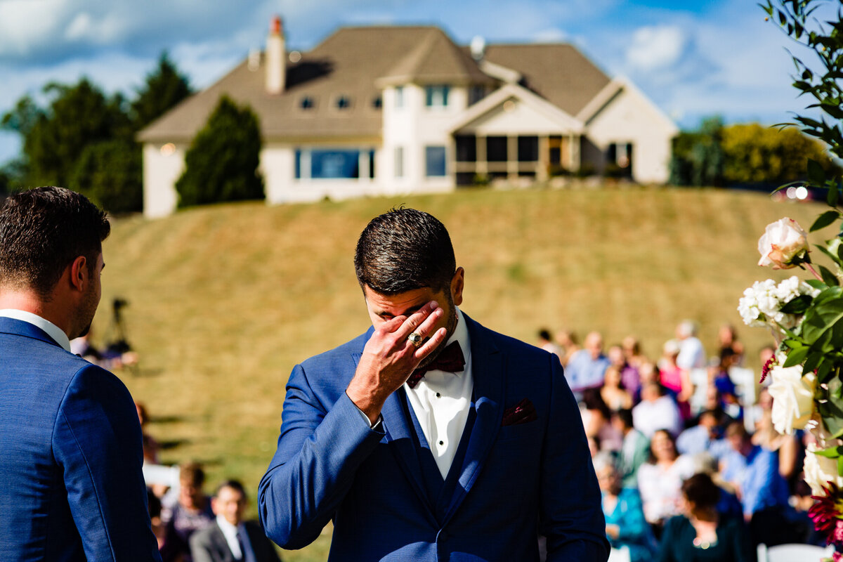 One of the top wedding photos of 2020. Taken by Adore Wedding Photography- Toledo, Ohio Wedding Photographers. This photo is of groom wiping tears from his eyes as the bride and her father walk down the aisle at summer outdoor wedding in Toledo Ohio