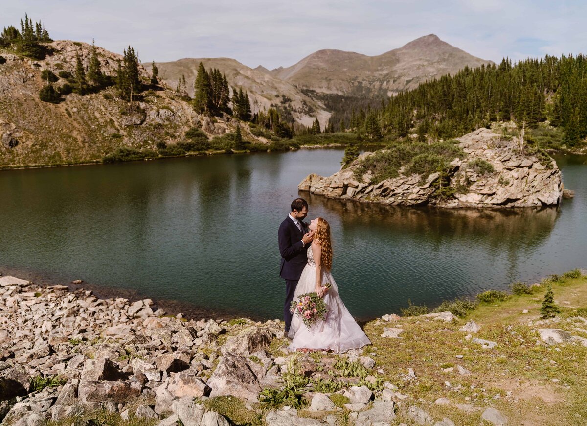 Bride and groom sharing a romantic moment on the rocky shore of an alpine lake during their Buena Vista, Colorado elopement, surrounded by mountains and pine trees.
