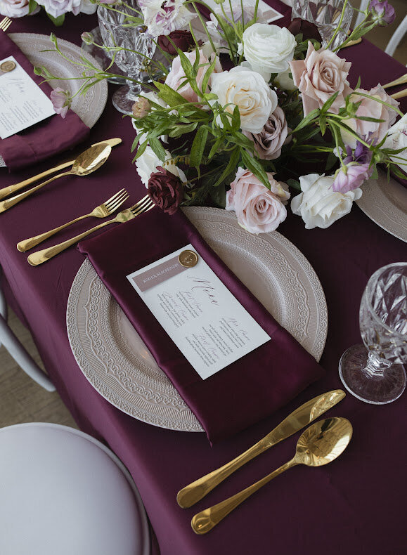 A close-up of an elegant burgundy and blush guest table at a National Arts Centre wedding, featuring a beautiful blush floral centrepiece and a place setting that includes an ornate blush charger plate layered with a burgundy satin napkin and a white menu with a blush place card and wax seal placed on the charger plate, surrounded by gold cutlery and crystal goblets.