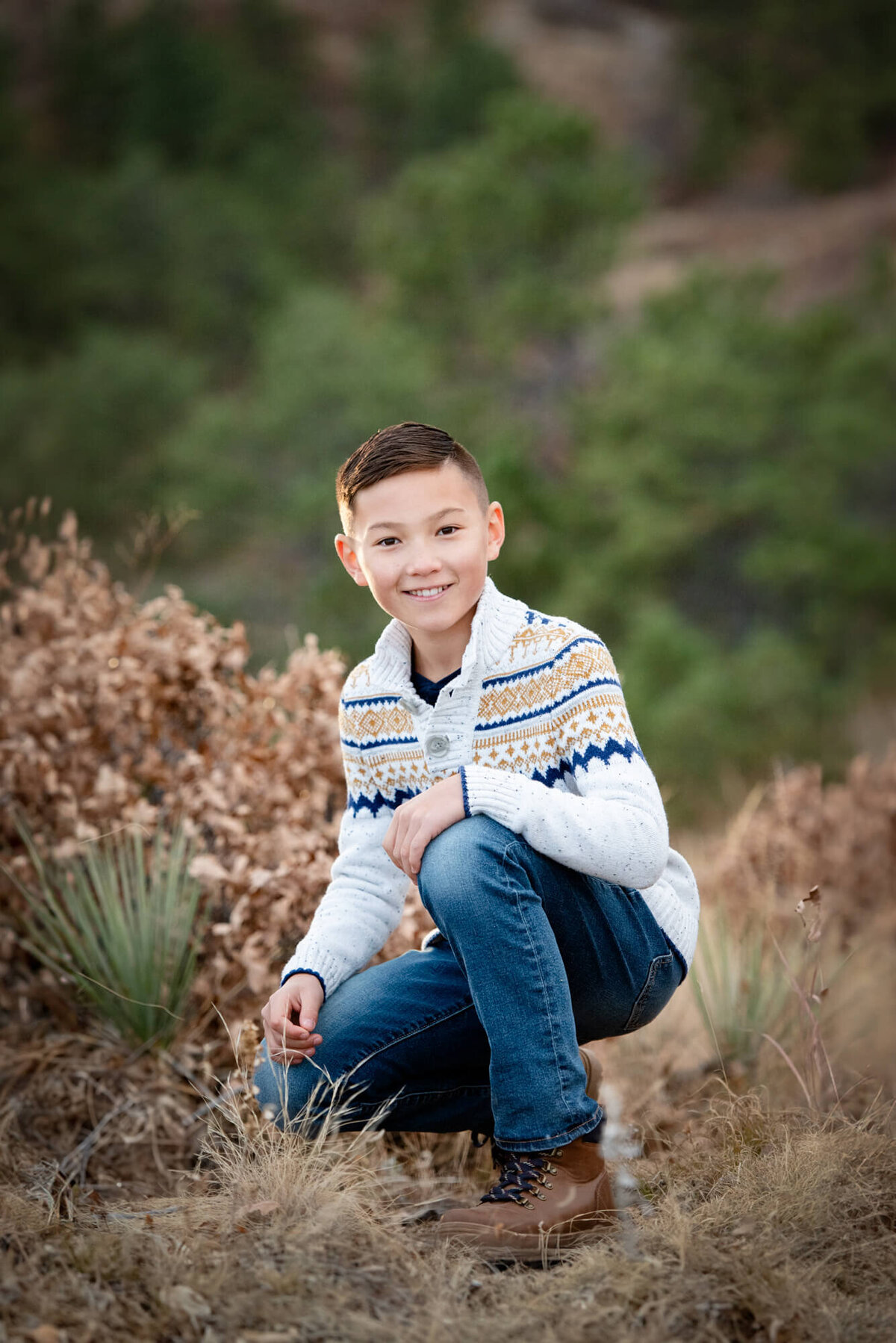 A young boy in a white sweater and jeans kneels in a mountain trail while smiling
