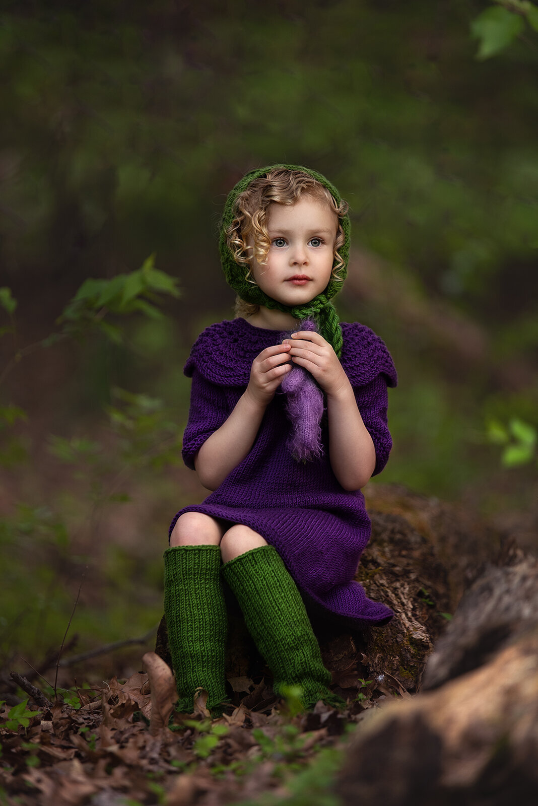 girl holding bear in purple dress and green bonnet and stockings outside on a log
