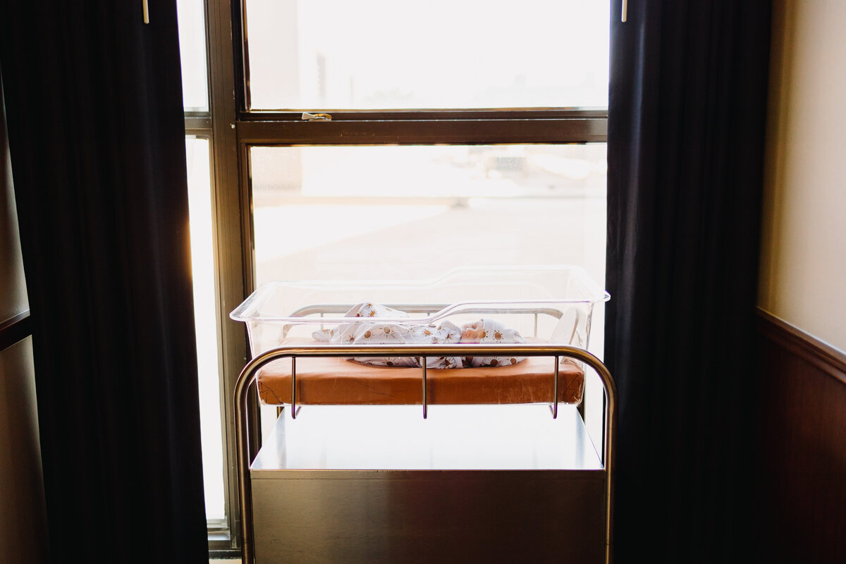 The artistry of Albuquerque's best Fresh 48 photographer with this serene newborn photograph. The baby, dressed in a beautiful pajama, peacefully sleeps in a white hospital cradle placed in front of a large window, capturing the gentle light and tranquility of the moment.