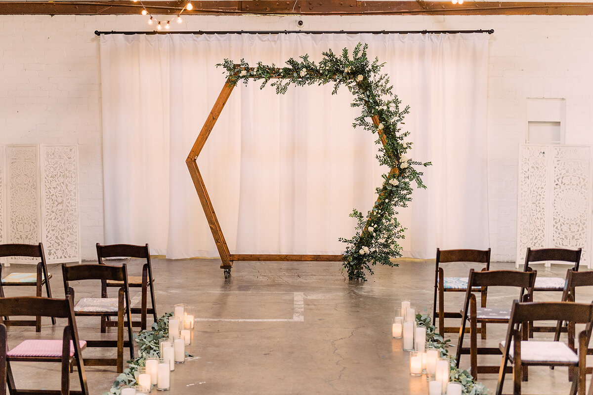 A minimalist hexagonal wooden arch decorated with greenery, placed in front of a white backdrop, with rows of chairs and a path lined with candles leading to it.