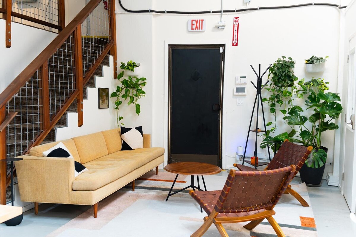 Cozy seating area featuring a mustard sofa, woven leather chairs, and abundant greenery near a stairwell.