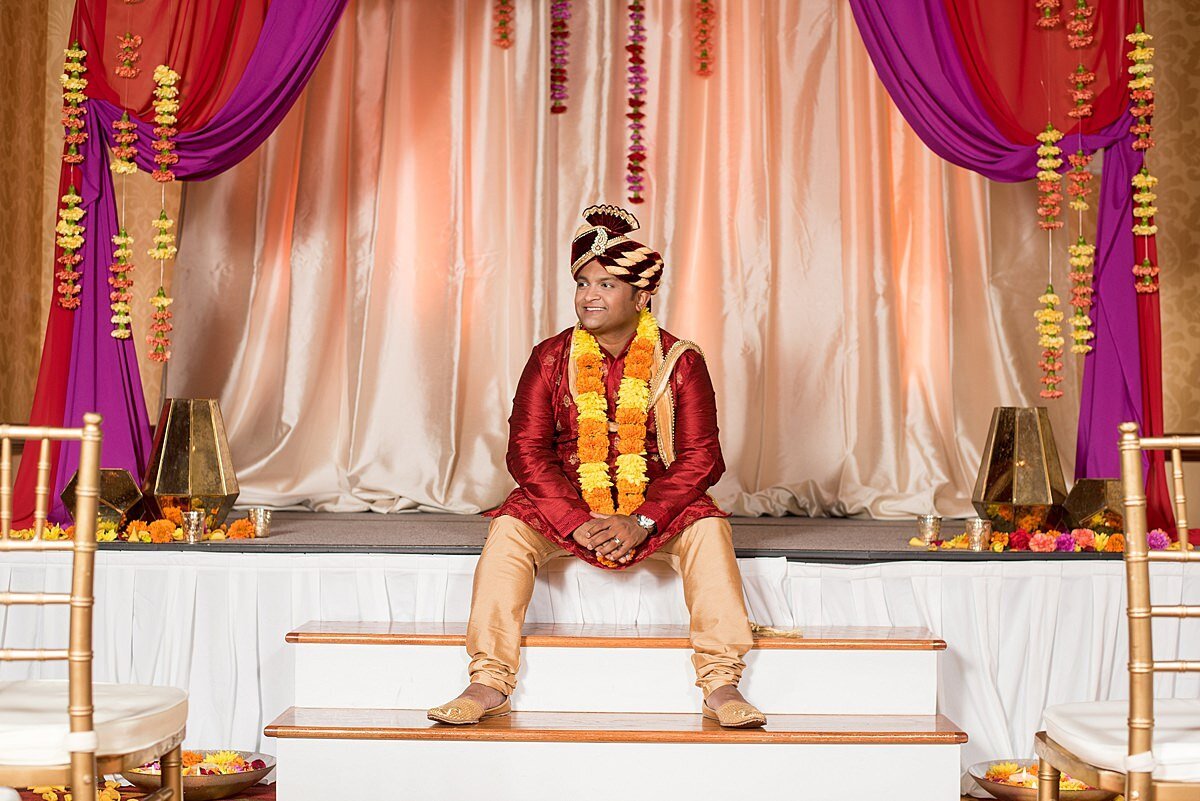 Hindu groom at Indian Wedding in Nashville sitting at the edge of the peach, magenta and red mandap wearing a red and gold sherwani with a yellow and orange varmala.