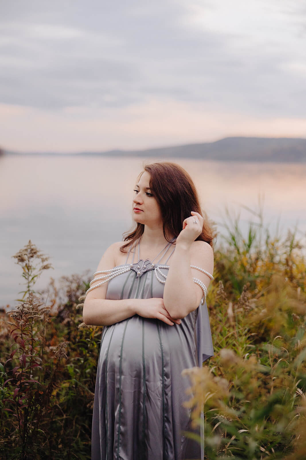 Pregnant woman wearing a silk dress standing by lake for maternity photo session in Fingerlakes, NY