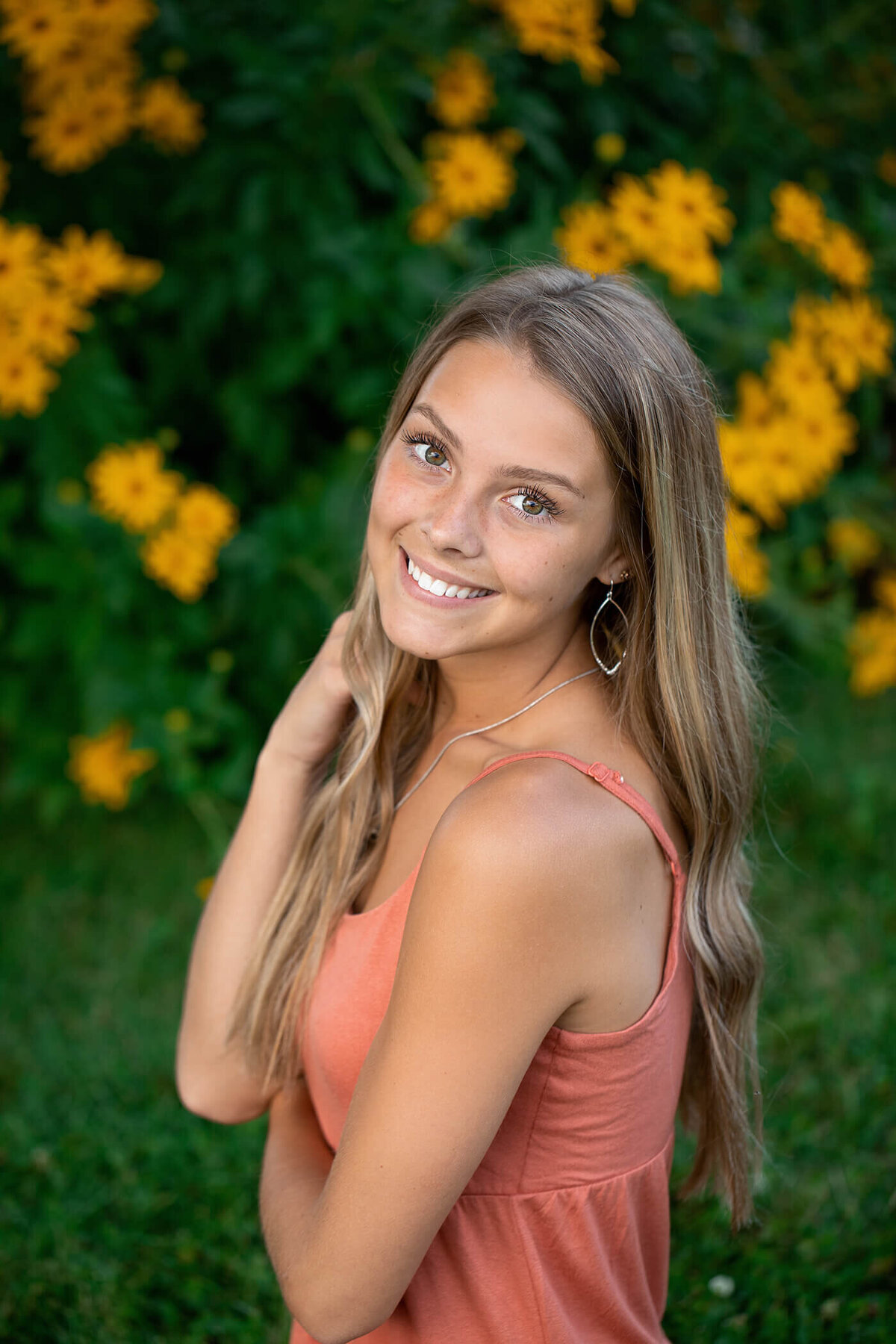 Senior girl in a peach top standing in front of a bunch of yellow flowers.