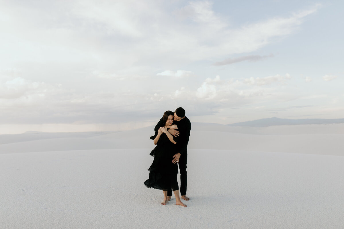 man holding fiancé from behind kissing her on a sand dune in New Mexico