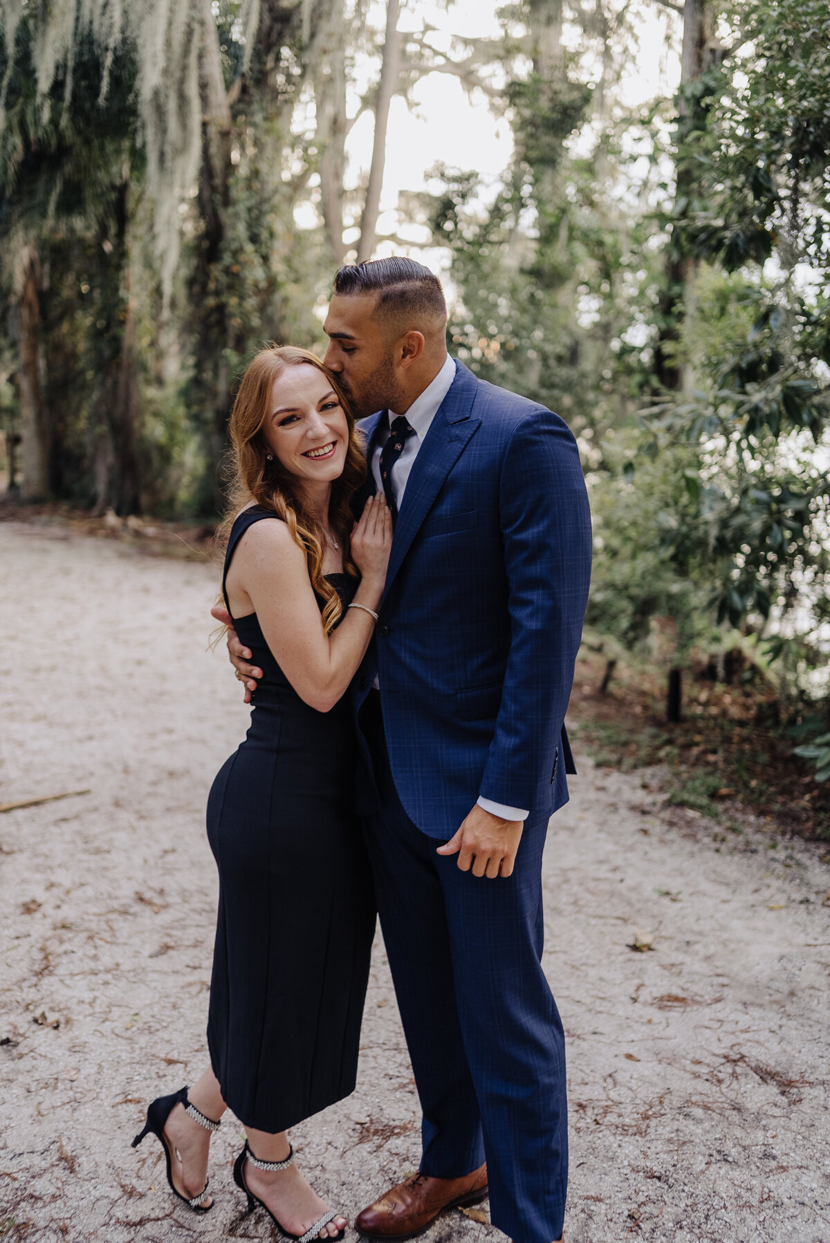 Engaged couple embrace. She has red hair and a navy dress; he wears a navy suit and leans down to kiss her on the temple. Photo taken by Orlando Wedding Photographer Four Loves Photo and Film.