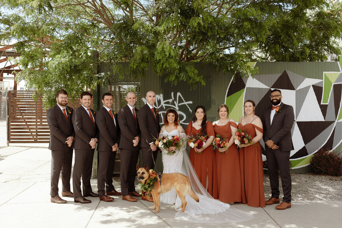 Bride and groom pose with their wedding party in black tuxes and orange bridesmaid dresses.