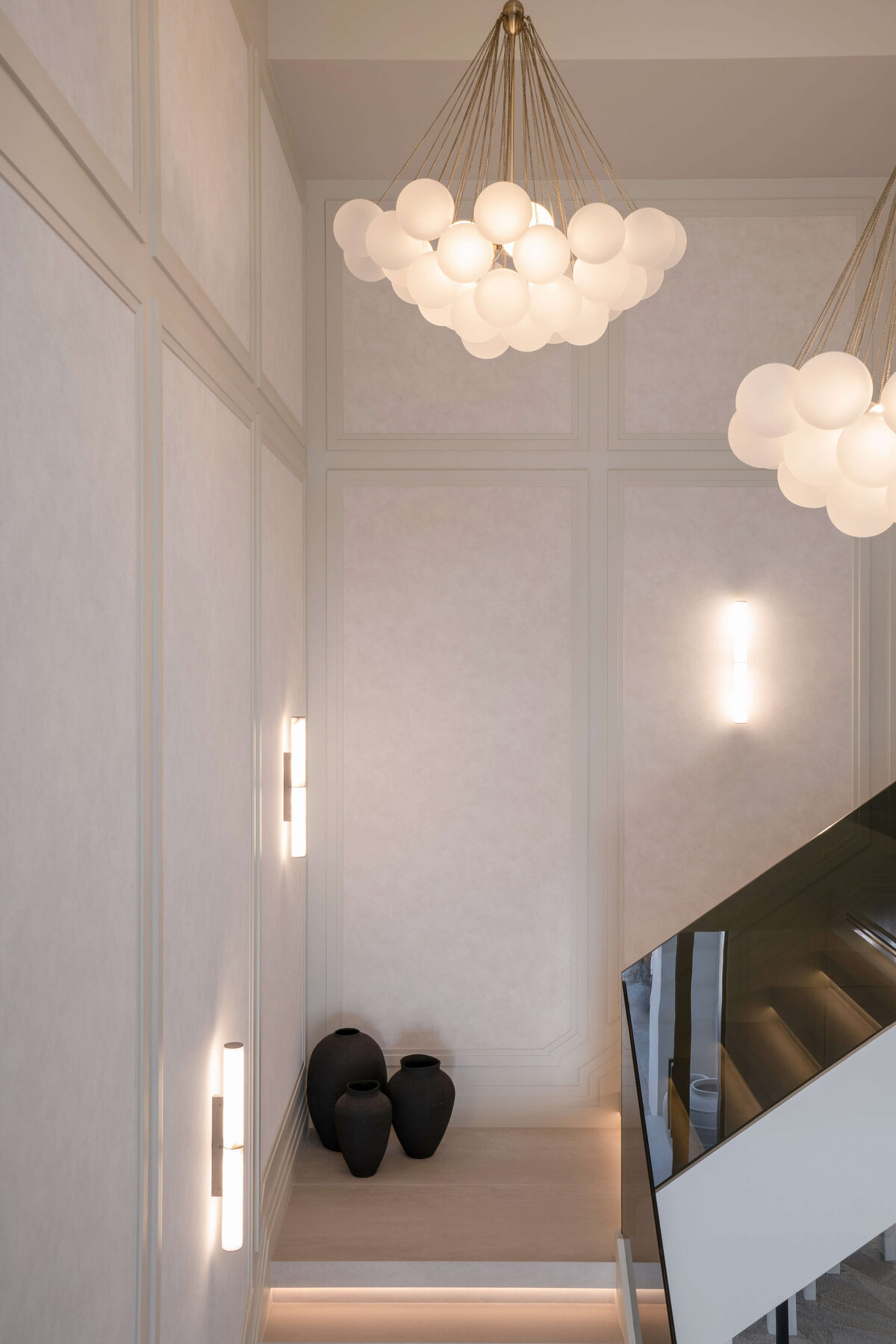 A modern staircase with a minimalist design featuring wide, white steps. The stairs are bordered by a sleek, black glass railing on the right side. The walls are adorned with vertical, cylindrical wall sconces that emit a warm glow, along with contemporary bubble chandeliers above. At the landing of the staircase, black decorative vases are visible, adding a touch of contrast to the light-colored interior.