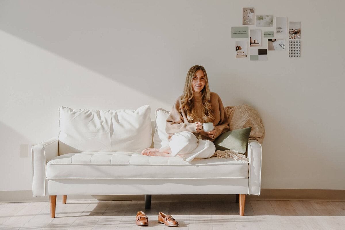 A woman sits on a white sofa in a bright room, smiling and holding a cup. She wears a beige sweater and white pants. A collage of small photos, showcasing creative web design ideas, decorates the wall above the sofa, while a pair of branded shoes rests on the floor.