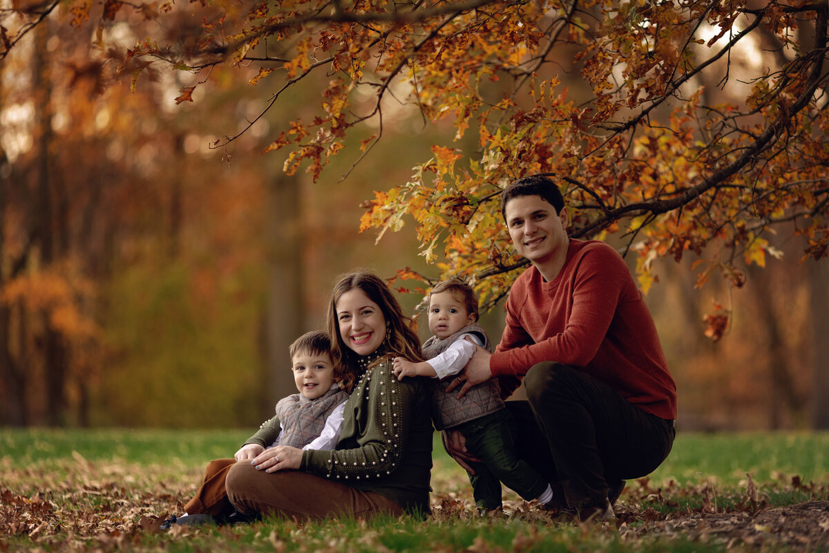 Happy parents sit in a park under a tree with their toddler son and daughter in their laps taken by a New Jersey Family Photographer