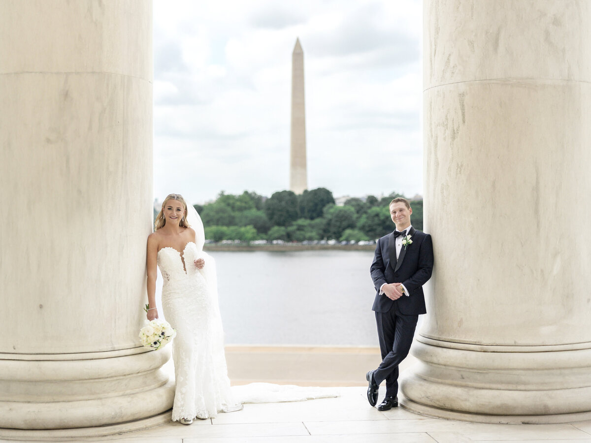 A bride and groom pose between marble columns, with the Washington Monument visible in the background. The bride holds a bouquet and wears a strapless white gown, while the groom is in a black suit with a boutonniere.