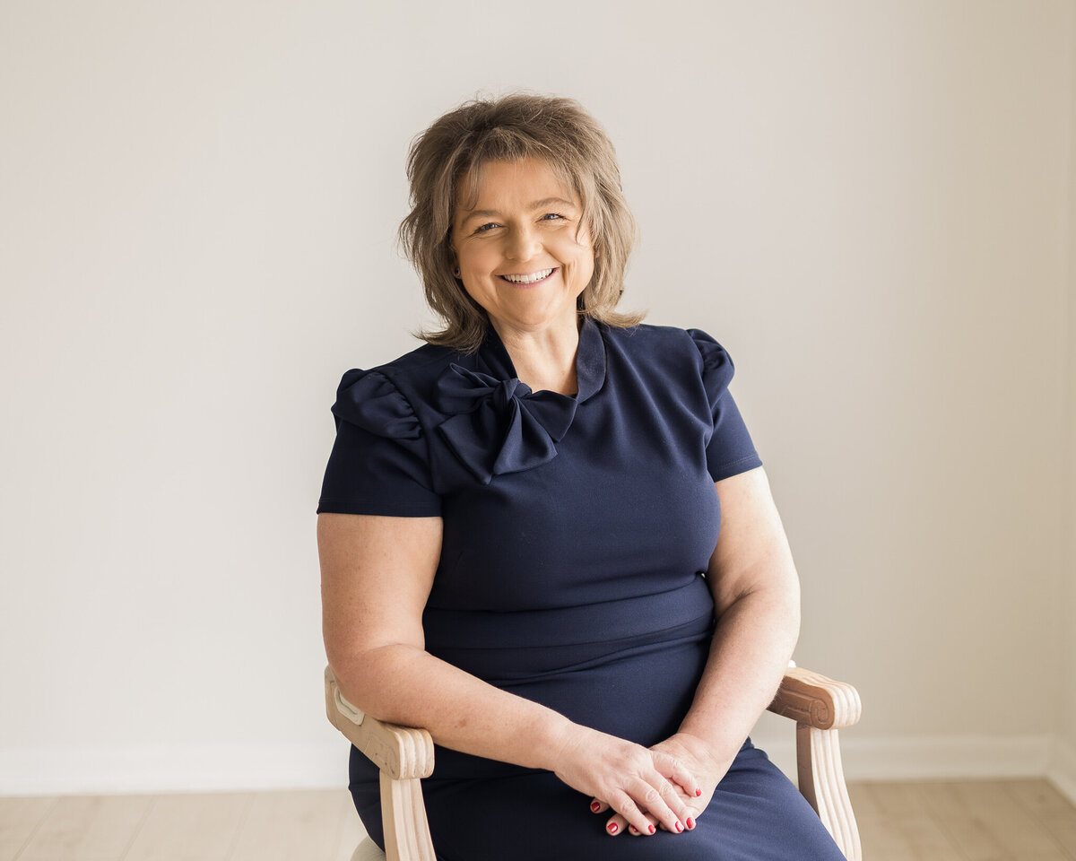 Brunette woman with shoulder length hair, sitting in a chair, wearing a dark navy dress, smiling at the camera
