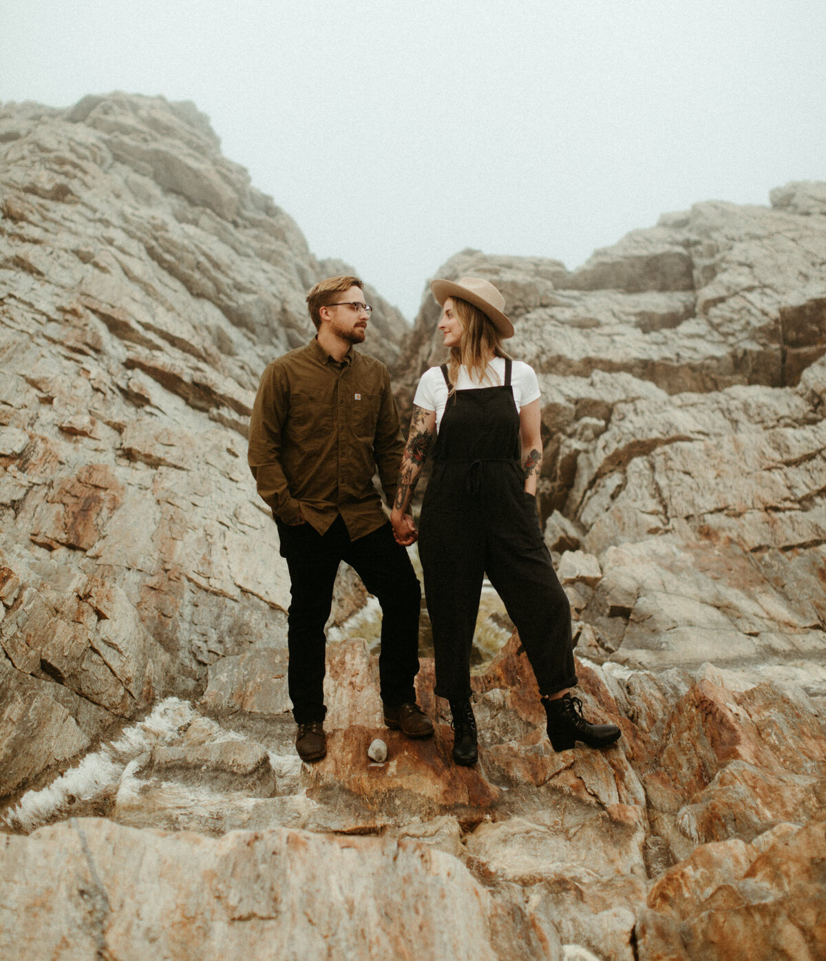 A girl wearing a wide brim hat, black overalls and boots holds hands with a guy wearing an olive green shirt and black pants as they stand on a rocky shoreline on a foggy day.