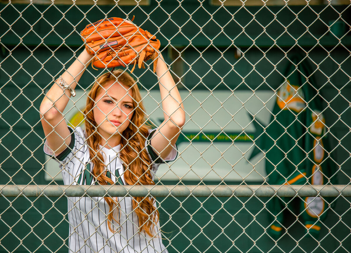 A high school graduate leans against a dugout fence on her elbows while wearing her glove.