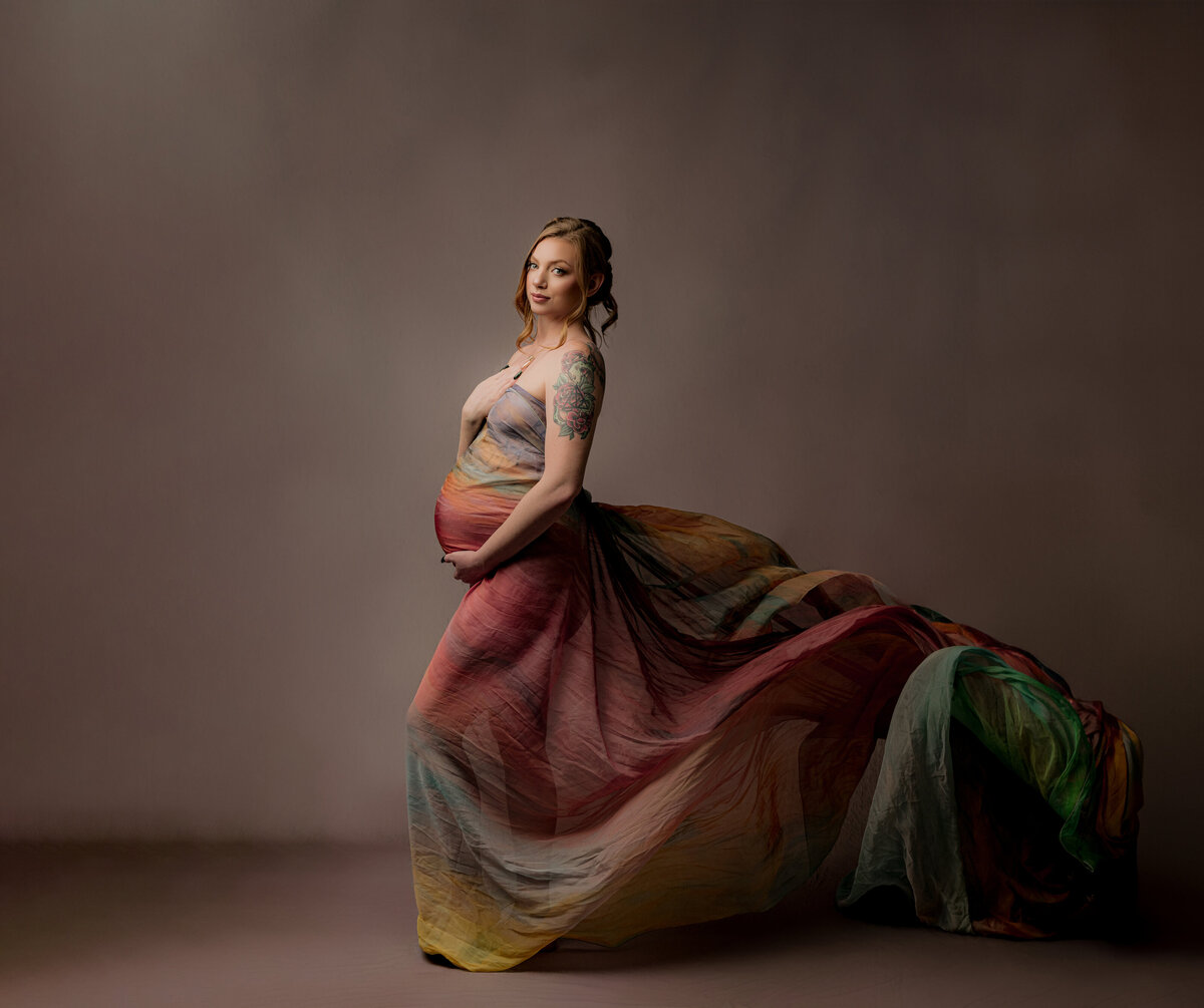 Pregnant mother holding belly in ombre rainbow fabric flowing looking straight at the camera  