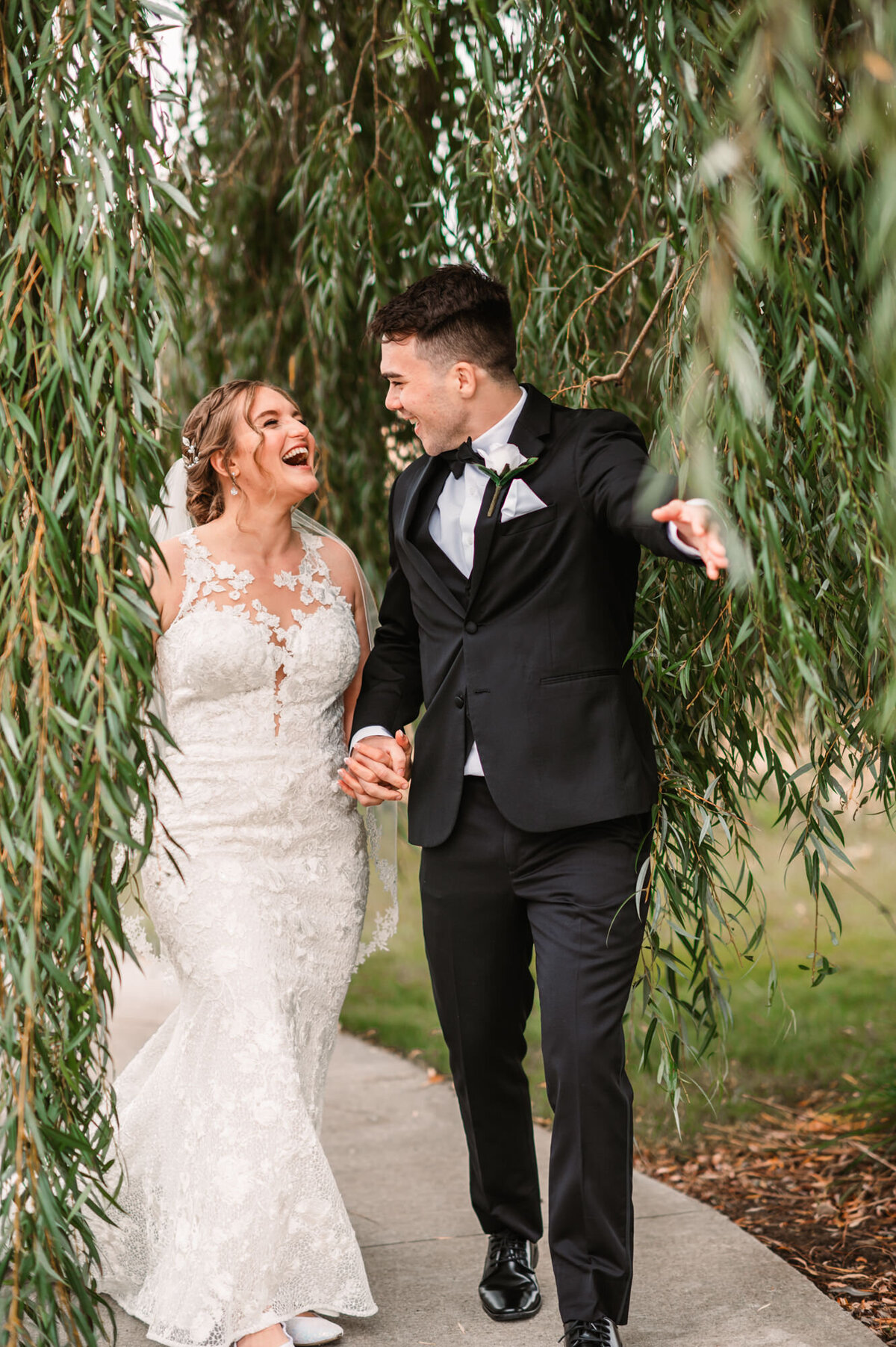 bride and groom laugh while holding hands and walking through a willow tree