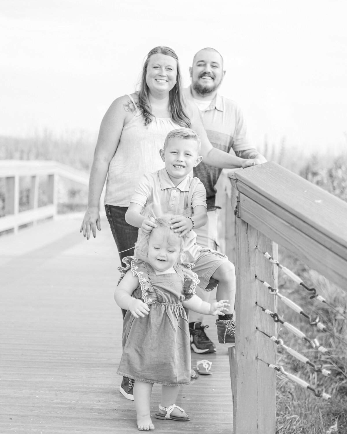 Black and white photo of mom, dad, young son, and toddler daughter on a bridge.  Mom and dad are looking at the camera smiling while brother has his hands on his sister's head, looking at the camera smiling.  Toddler girl is looking down smiling.