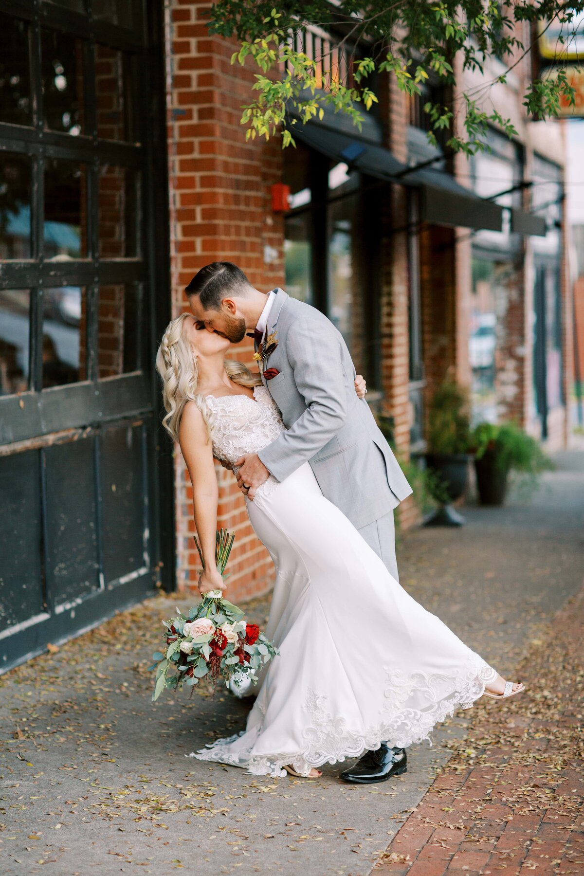 Bride and groom share romantic dip kiss outside on the street with the bride leaning back holding her bouquet of orchids and roses.