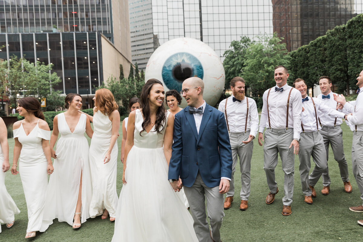 An outdoor portrait of a bride and groom walking with their wedding party in front of the iconic Giant Eyeball at The Joule hotel in Dallas, Texas.  The bride is on the left and is wearing a sleeveless, white dress. The groom is on the right and is wearing a blue jacket, blue bowtie, grey pants, and glasses. They are both holding hands and smiling at each other. The wedding party are either wearing white dresses or white dress shirts with grey pants, suspenders, and bowties.