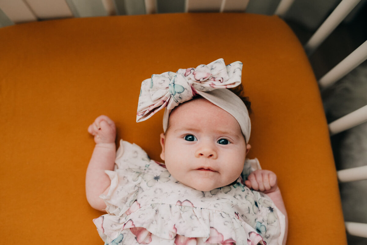 A baby girl wearing a soft pink outfit and matching bow looks right at the camera during her photoshoot with Allison Wolf, Boston Newborn Photographer