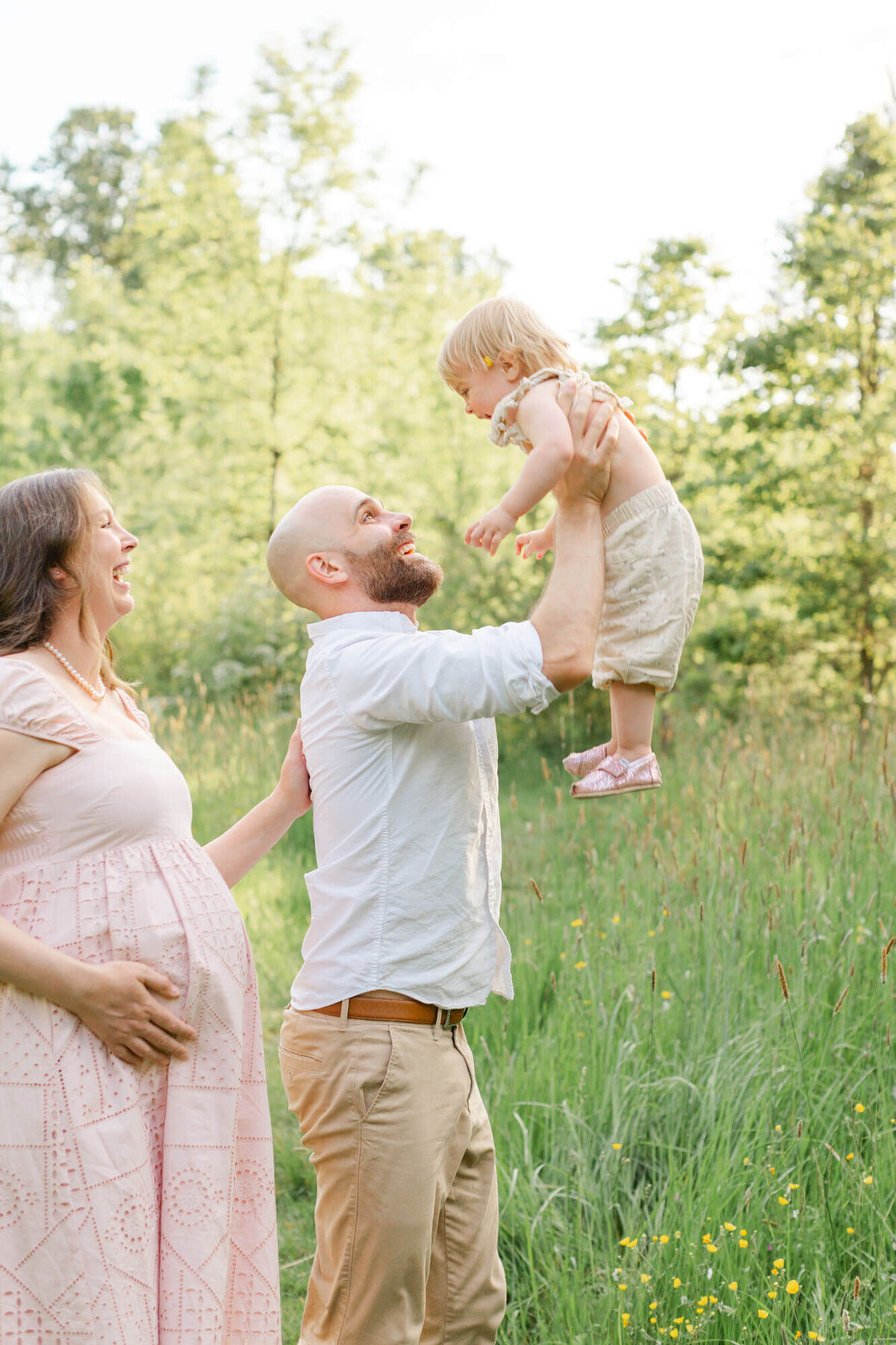 Dad is holding toddler up in the air like he is going to throw her up and pregnant mom is standing behind dad watching. Everyone has big smiles on their faces. They are all wearing neutral colors, mom is in a light pink dress.