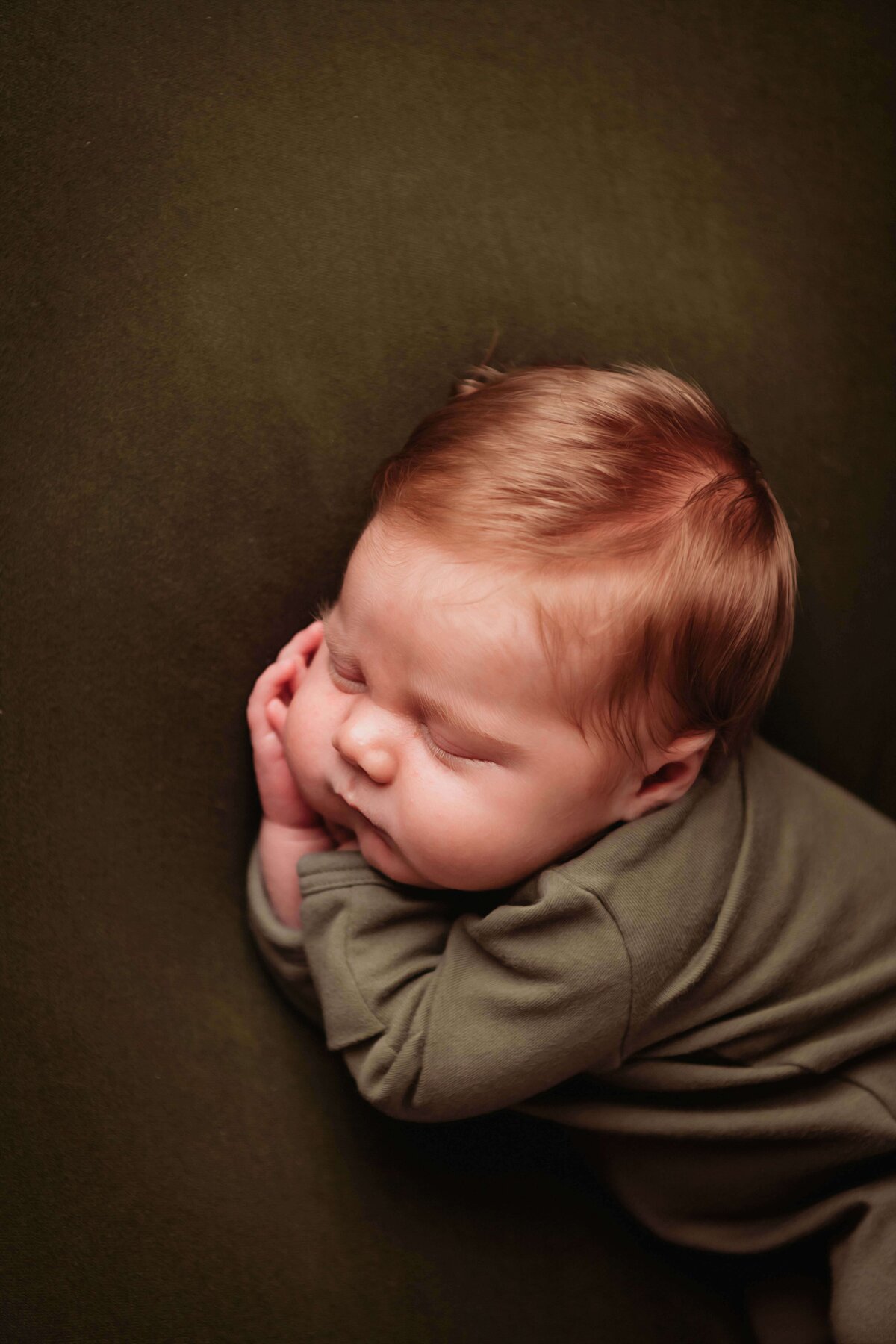 A red headed newborn baby boy, sleeping peacefully in green pajamas on a matching green blanket. He has his hands tucked gently under his chubby cheeks, as he sleeps in this gorgeous newborn photo by a Mobile, AL newborn photographer