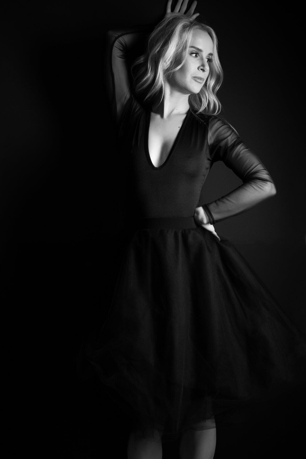 Elegant black-and-white portrait of a woman in a black dress with sheer sleeves and a tulle skirt, striking a graceful pose against a dark background.