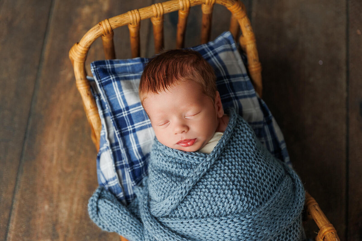 A peaceful newborn is swaddled in a soft blue knitted blanket, lying in a wicker basket. The basket is placed on a plaid pillow, creating a cozy scene. The baby has closed eyes and a faint smile, with short brown hair visible.