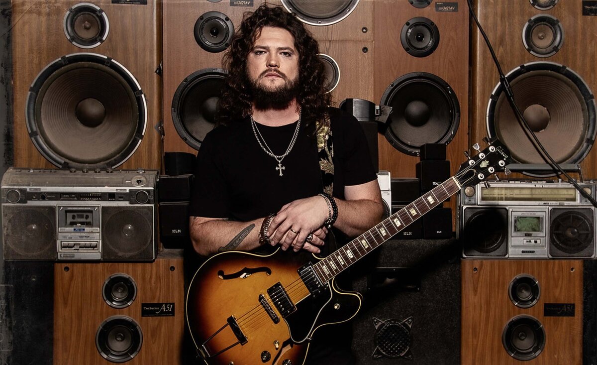 Male musician photo Daniel Martin wearing black t shirt holding sunburst electric guitar with speaker wall behind