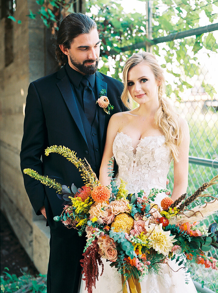 Bride and groom, wearing a black tuxedo and white wedding gown, pose with a large bouquet of yellow, blush and orange flowers.