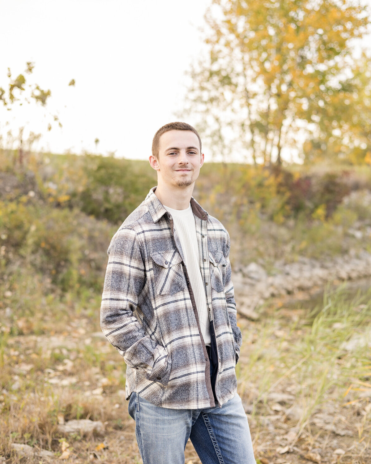 high school senior male standing at an angle next to a pond, wearing a blue flannel jacket with his hands in his pocket, blue jeans, light tan shirt, and smiling softly at the camera