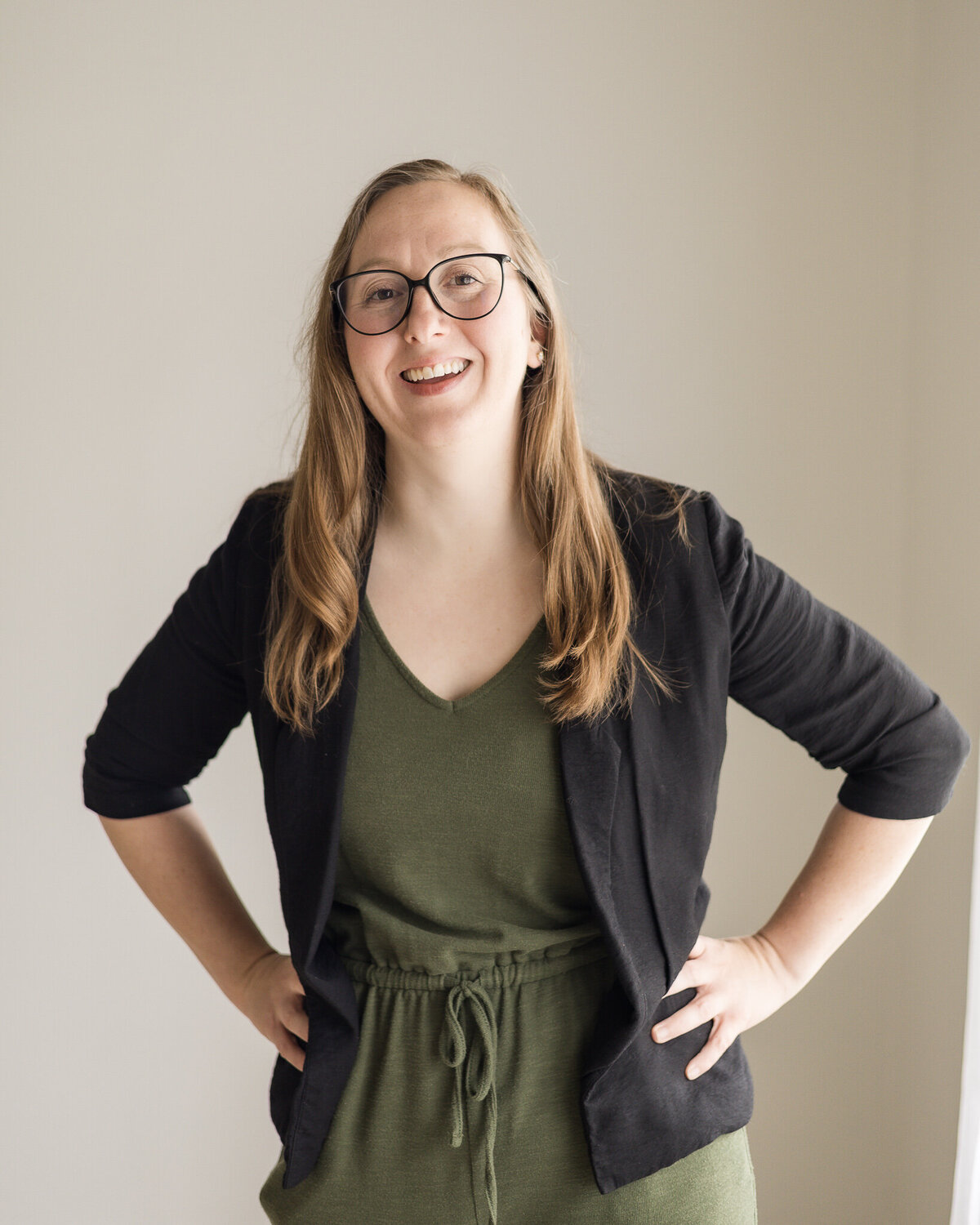 Strawberry blonde female, standing with her hands on her hips, wearing a dark green shirt and pants, with a black jacket over the green outfit, wearing glasses, smiling at the camera