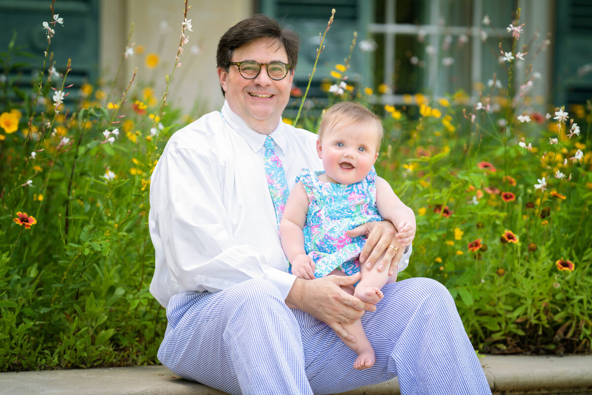 Dad and baby daughter are sitting in some wildflowers at Longue Vue House and Gardens.  Dad is wearing seersucker pants and a white dress shirt.  He has short, dark hair and glasses.  Baby is wearing Lily Pulitzer and sitting on his lap smiling at the camera.  She has blonde hair.