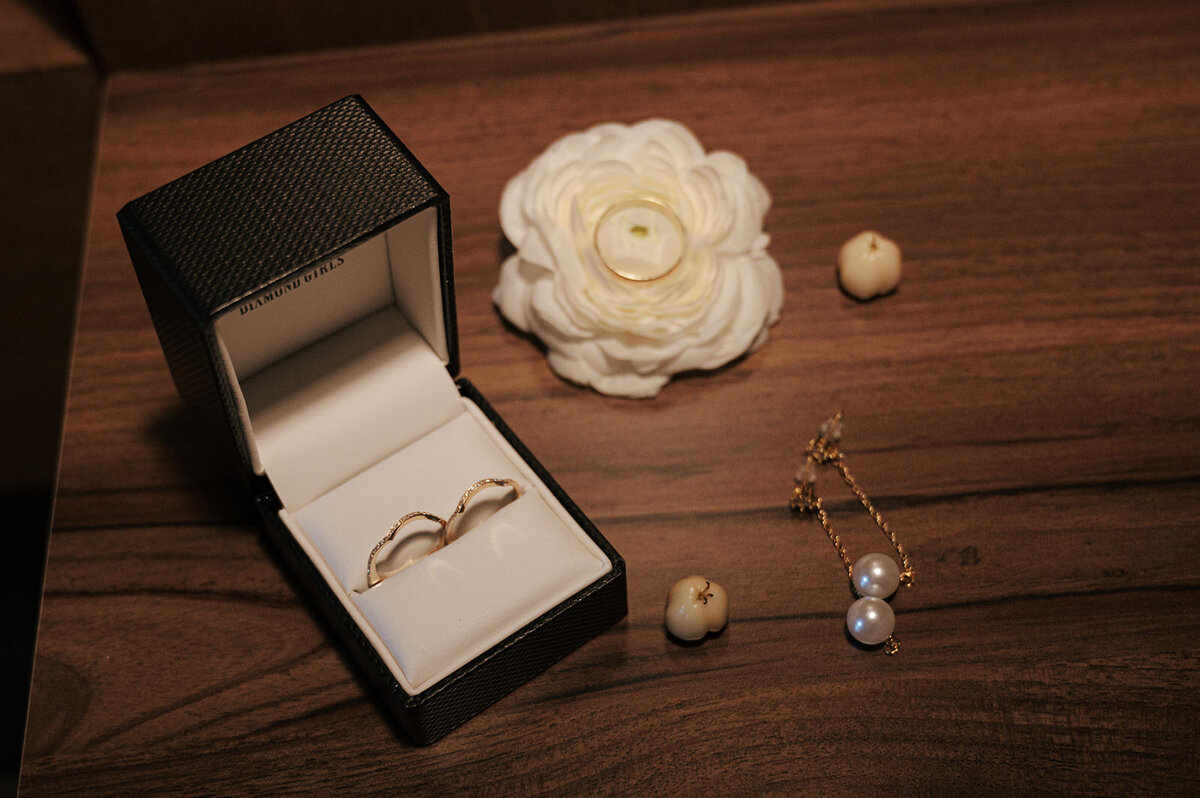 A jewelry box containing two rings sits on a wooden surface along with a pair of pearl earrings.