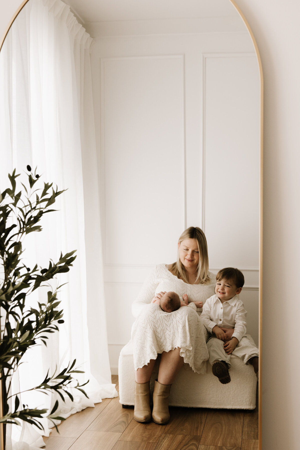 A mother sits in a softly lit studio, cradling her newborn in her lap while her toddler smiles beside her. Captured through a mirror, the moment is framed by sheer curtains and an olive tree, creating a peaceful and elegant scene.