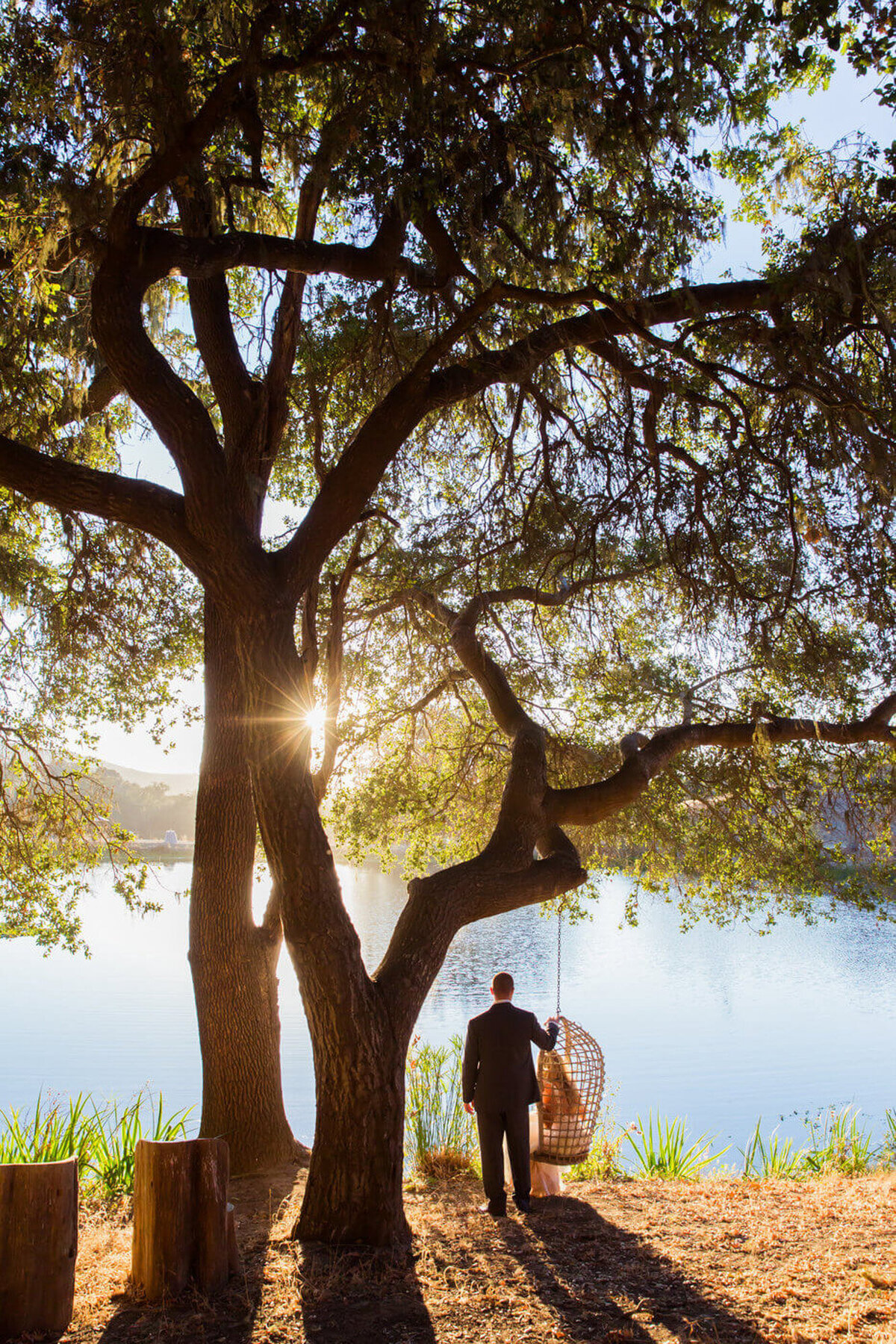 Sunset view at the lakeside while bride sitting on a swing hanging from the tree, while groom is beside her